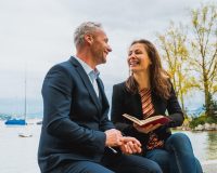 Coppia adulta che sorride mentre legge insieme un libro ad alta voce all’aperto, vicino all’acqua.